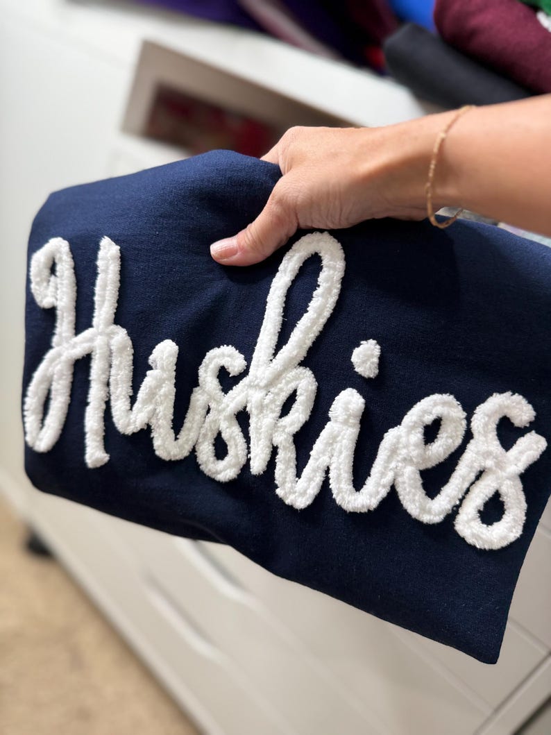 May include: A navy blue sweatshirt with the word "Huskies" in large, white, textured lettering. The sweatshirt is held up, showcasing the design. The background is blurred, with other folded garments visible.