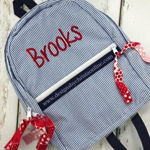 May include: Blue and white striped backpack with the name "Brooks" embroidered in red. The backpack has a white zipper, navy blue straps, and red and white patterned ribbons attached. The website address www.designsbychristaonline.com is printed on the front.