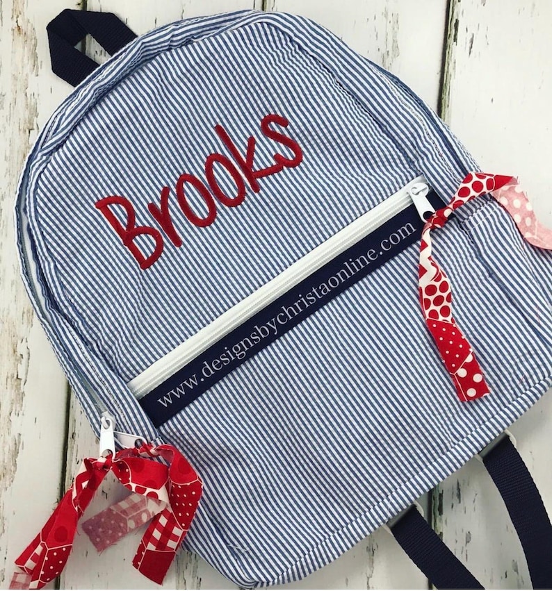 May include: Blue and white striped backpack with the name "Brooks" embroidered in red. The backpack has a white zipper, navy blue straps, and red and white patterned ribbons attached. The website address www.designsbychristaonline.com is printed on the front.