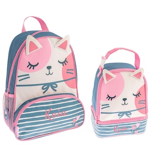 May include: A blue and pink cat-themed backpack and lunch bag set. The backpack has a large cat face with pink ears and a pink bow. The lunch bag has a smaller cat face with pink ears and a pink bow. Both items have a blue and white striped pattern and the name "Kaia" is printed on the bottom.