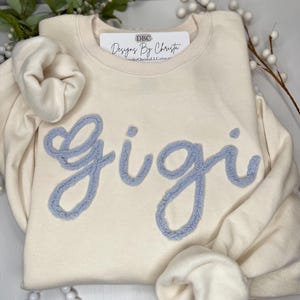 May include: A cream coloured sweatshirt with a light blue fuzzy appliqu&eacute; that says "Gigi". The sweatshirt has a tag that says "DBC Designs By Christa".