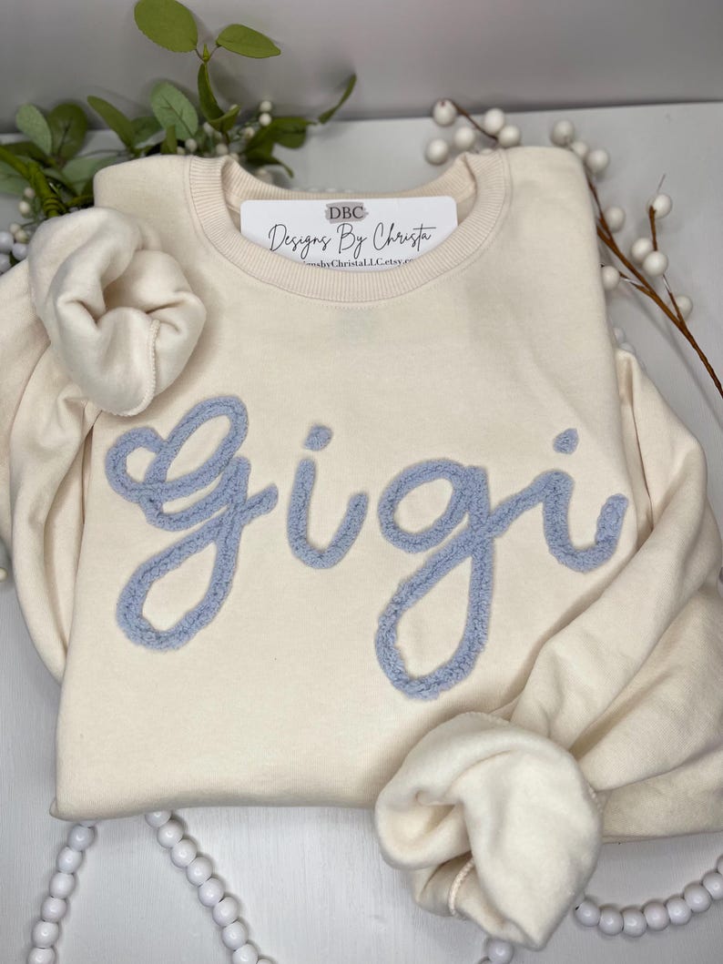 May include: A cream coloured sweatshirt with a light blue fuzzy appliqu&eacute; that says "Gigi". The sweatshirt has a tag that says "DBC Designs By Christa".