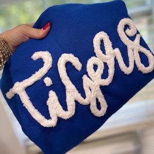 May include: Royal blue sweatshirt with the word "Tigers" in white, textured, cursive lettering. The sweatshirt is held up by a hand with red nail polish and gold bracelets.