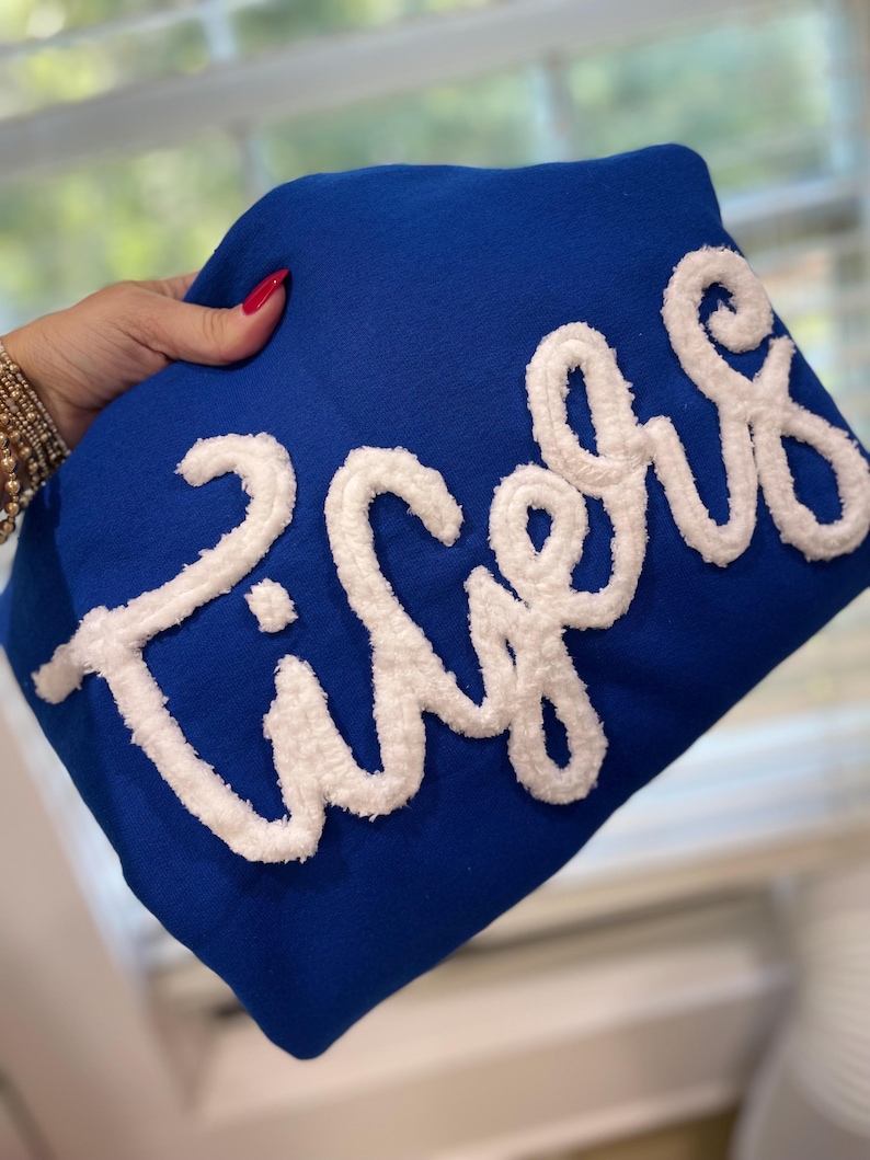 May include: Royal blue sweatshirt with the word "Tigers" in white, textured, cursive lettering. The sweatshirt is held up by a hand with red nail polish and gold bracelets.