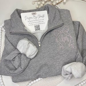 May include: Gray zippered sweatshirt with a pink bow and the name "Lmm" embroidered on the front. The sweatshirt is folded and laid on a white surface.