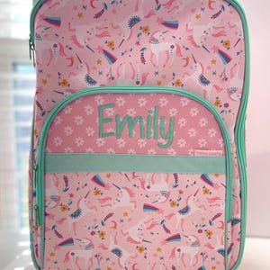 May include: Pink rolling suitcase with a unicorn and floral pattern. The suitcase has a green trim and wheels. The name "Emily" is embroidered on the front of the suitcase.