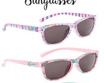 May include: Three pairs of colorful, personalized sunglasses for kids. The sunglasses have different patterns and designs, including stripes, polka dots, and flowers. Each pair has a name on the side: "Chip", "Kaia", and "Anna".
