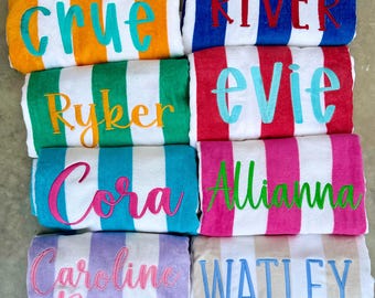 May include: A collection of personalized beach towels with vertical stripes in various colors, including orange, blue, green, red, pink, and purple. Each towel features an embroidered name in a contrasting color.