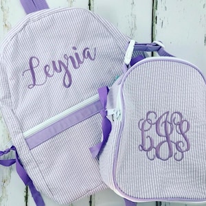 May include: A matching backpack and lunch bag set. Both are light purple and white striped with purple accents. The backpack has the name "Leyria" embroidered on it. The lunch bag has a monogram.