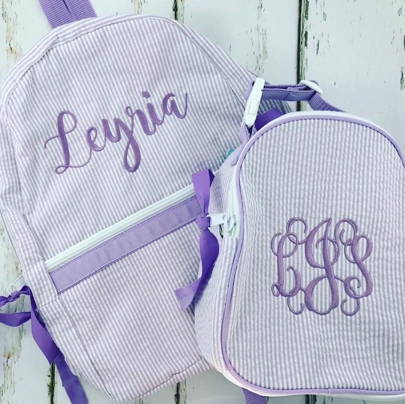 May include: A matching backpack and lunch bag set. Both are light purple and white striped with purple accents. The backpack has the name "Leyria" embroidered on it. The lunch bag has a monogram.
