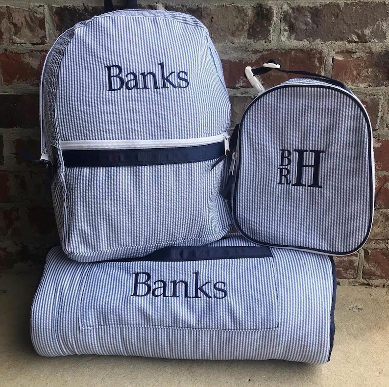 May include: A set of blue and white striped seersucker bags and a blanket. The backpack and blanket are embroidered with the name "Banks". The lunch bag has the initials "BRH".