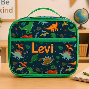 Może przedstawiać: Granatowa torba na lunch z zielonym uchwytem i wykończeniem. Torba ma wz&oacute;r dinozaur&oacute;w i imię "Levi" pomarańczowymi literami. Torba jest przeznaczona do przenoszenia jedzenia i napoj&oacute;w.