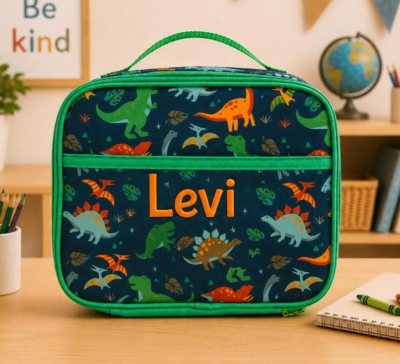 Może przedstawiać: Granatowa torba na lunch z zielonym uchwytem i wykończeniem. Torba ma wz&oacute;r dinozaur&oacute;w i imię "Levi" pomarańczowymi literami. Torba jest przeznaczona do przenoszenia jedzenia i napoj&oacute;w.