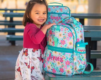 May include: A light blue backpack and matching lunch bag featuring a colorful floral pattern with pink, yellow, and green flowers. Both items are personalized with the name Anna in pink script. A matching light blue water bottle is included.
