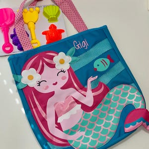 May include: A blue and pink tote bag with a mermaid design. The mermaid has pink hair, a pink shell bra, and a green tail. The bag has a mesh pocket and the name "Gigi" is written on the front.