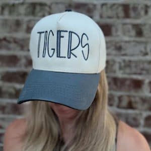 Op de afbeelding: Een cr&egrave;mekleurige baseballcap met een grijze klep. Het woord "TIGERS" is in marineblauw op de voorkant geborduurd. De cap is een klassieke stijl, geschikt voor vrijetijdskleding.