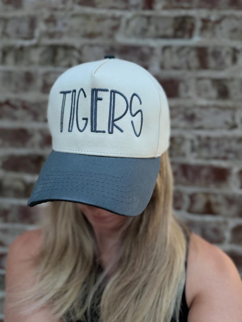 Op de afbeelding: Een cr&egrave;mekleurige baseballcap met een grijze klep. Het woord "TIGERS" is in marineblauw op de voorkant geborduurd. De cap is een klassieke stijl, geschikt voor vrijetijdskleding.
