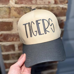 Op de afbeelding: Een beige en grijze baseballcap met het woord "TIGERS" in donkergrijs geborduurd. De cap heeft een gebogen klep en een gestructureerd ontwerp, voor sportfans.