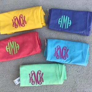 May include: Five folded t-shirts with different colors and monograms. The monograms are in pink, green, and light blue. The t-shirts are folded and stacked on a light gray surface.