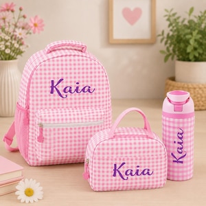 May include: A pink and white gingham backpack, lunch bag, and water bottle set. The backpack has a front pocket and side mesh pocket. The lunch bag has a handle. Each item is personalized with the name "Kaia" in purple script.