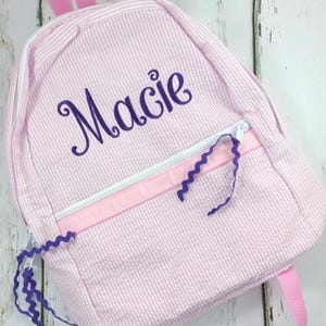 May include: Pink and white striped backpack with the name "Macie" embroidered in purple script. The backpack features a white zipper, pink ribbon accents, and purple zigzag ribbon details. Pink straps complete the design.