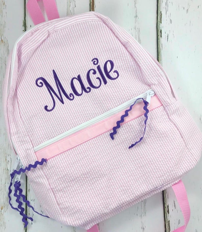 May include: Pink and white striped backpack with the name "Macie" embroidered in purple script. The backpack features a white zipper, pink ribbon accents, and purple zigzag ribbon details. Pink straps complete the design.