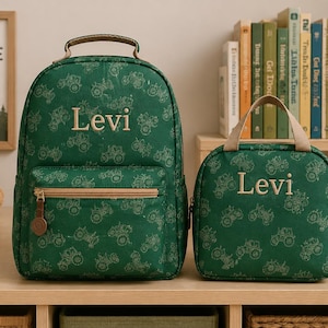May include: A green backpack and a matching lunch bag, both featuring a repeating tractor pattern and the name Levi embroidered in light beige. Each bag has a top handle, a front zippered pocket, and light beige trim accents.
