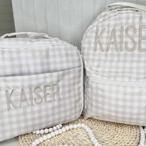 May include: A matching set of beige and white gingham patterned backpack and lunch bag. Both bags have the name "KAISER" embroidered in a matching beige thread. The backpack has a front pocket and a top handle.