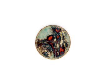 Broche redondo de cobre y esmalte hecho a mano: joyería única con diseños abstractos en turquesa y rojo.