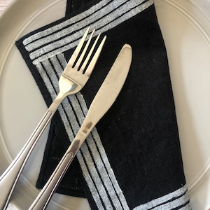 Puede incluir: Una servilleta de lino negra con un borde bordado blanco. La servilleta está doblada y colocada en un plato blanco con un tenedor y un cuchillo de plata.