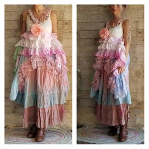 K&ouml;nnte beinhalten: Ein langes, mehrschichtiges Kleid mit einem wei&szlig;en Spitzenoberteil und einer Kaskade von R&uuml;schen in Pastellt&ouml;nen. Das Kleid hat eine gro&szlig;e rosa Blume und ein flie&szlig;endes, b&ouml;hmisches Design. Es ist ein einzigartiges, k&uuml;nstlerisches Kleidungsst&uuml;ck.