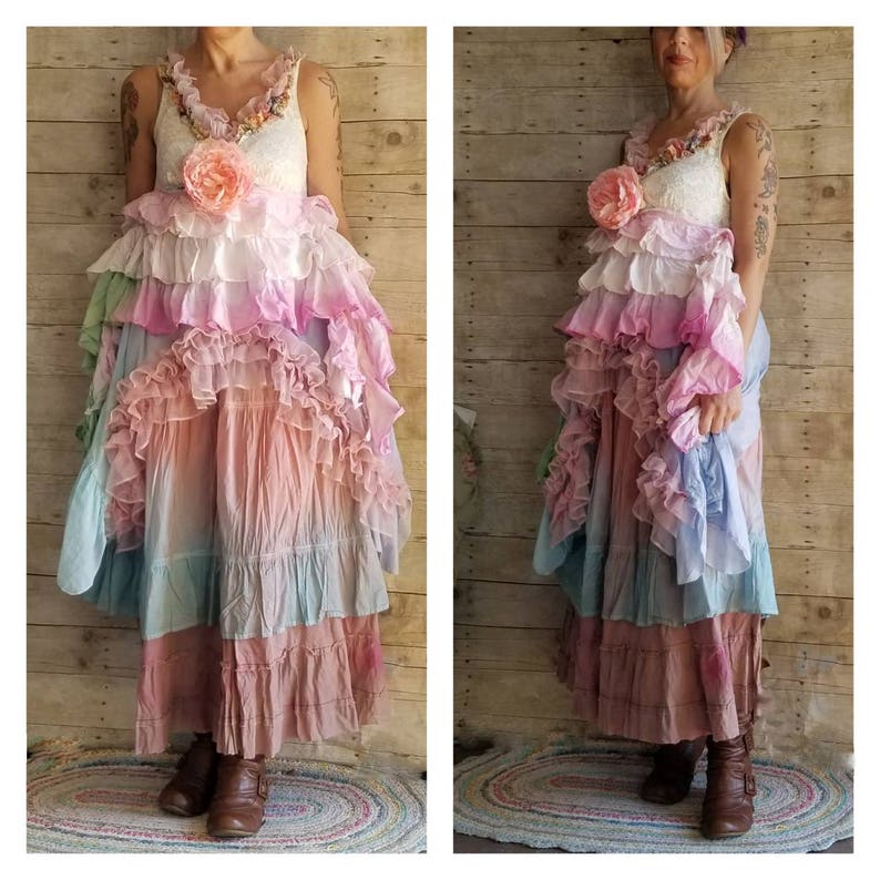K&ouml;nnte beinhalten: Ein langes, mehrschichtiges Kleid mit einem wei&szlig;en Spitzenoberteil und einer Kaskade von R&uuml;schen in Pastellt&ouml;nen. Das Kleid hat eine gro&szlig;e rosa Blume und ein flie&szlig;endes, b&ouml;hmisches Design. Es ist ein einzigartiges, k&uuml;nstlerisches Kleidungsst&uuml;ck.