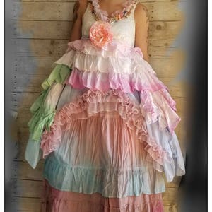K&ouml;nnte beinhalten: Ein mehrschichtiges Kleid in Pastellt&ouml;nen mit einem wei&szlig;en Spitzenoberteil, verziert mit einer rosa Blume und R&uuml;schen. Der Rock hat Schichten aus Rosa, Blau und Gr&uuml;n. Die Person tr&auml;gt braune Stiefel. Boho-Stil.