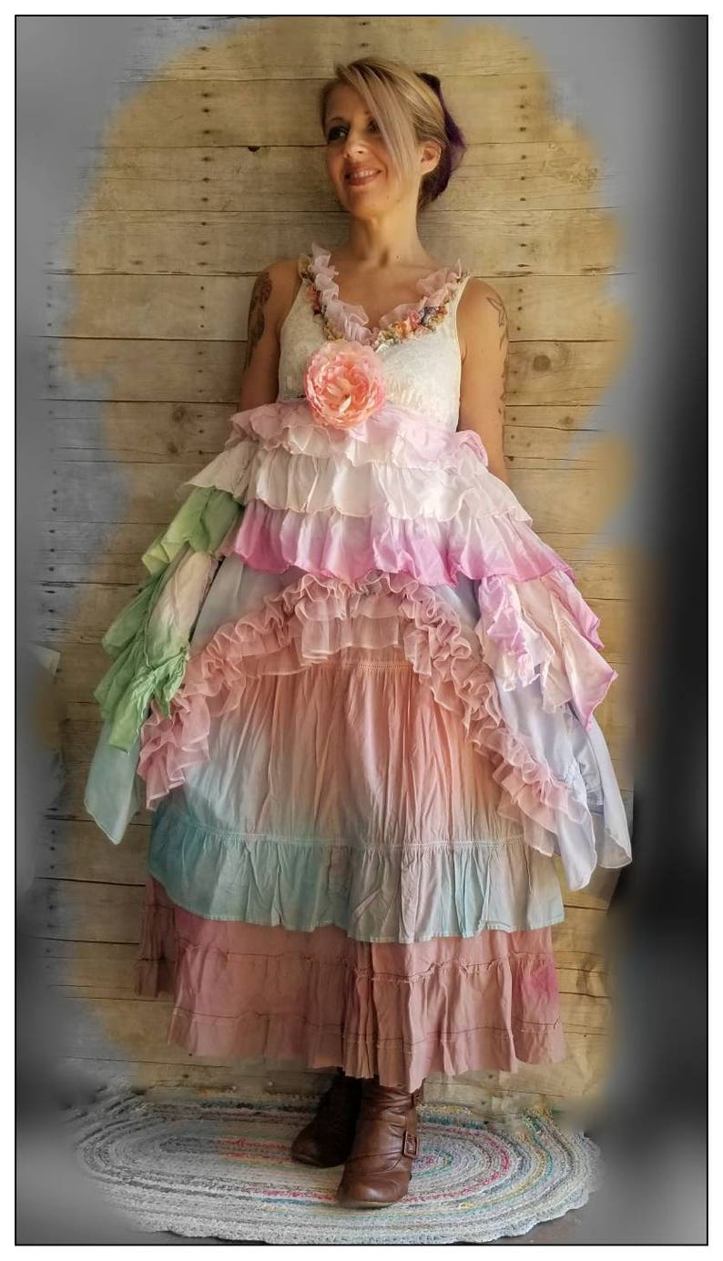 K&ouml;nnte beinhalten: Ein mehrschichtiges Kleid in Pastellt&ouml;nen mit einem wei&szlig;en Spitzenoberteil, verziert mit einer rosa Blume und R&uuml;schen. Der Rock hat Schichten aus Rosa, Blau und Gr&uuml;n. Die Person tr&auml;gt braune Stiefel. Boho-Stil.