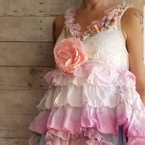 K&ouml;nnte beinhalten: Ein mehrlagiges R&uuml;schenkleid in Rosa-, Wei&szlig;- und Blaut&ouml;nen. Das Kleid hat eine gro&szlig;e, rosa Stoffblume am Oberteil und zarte Spitzendetails. Der Gesamtstil ist romantisch und b&ouml;hmisch.