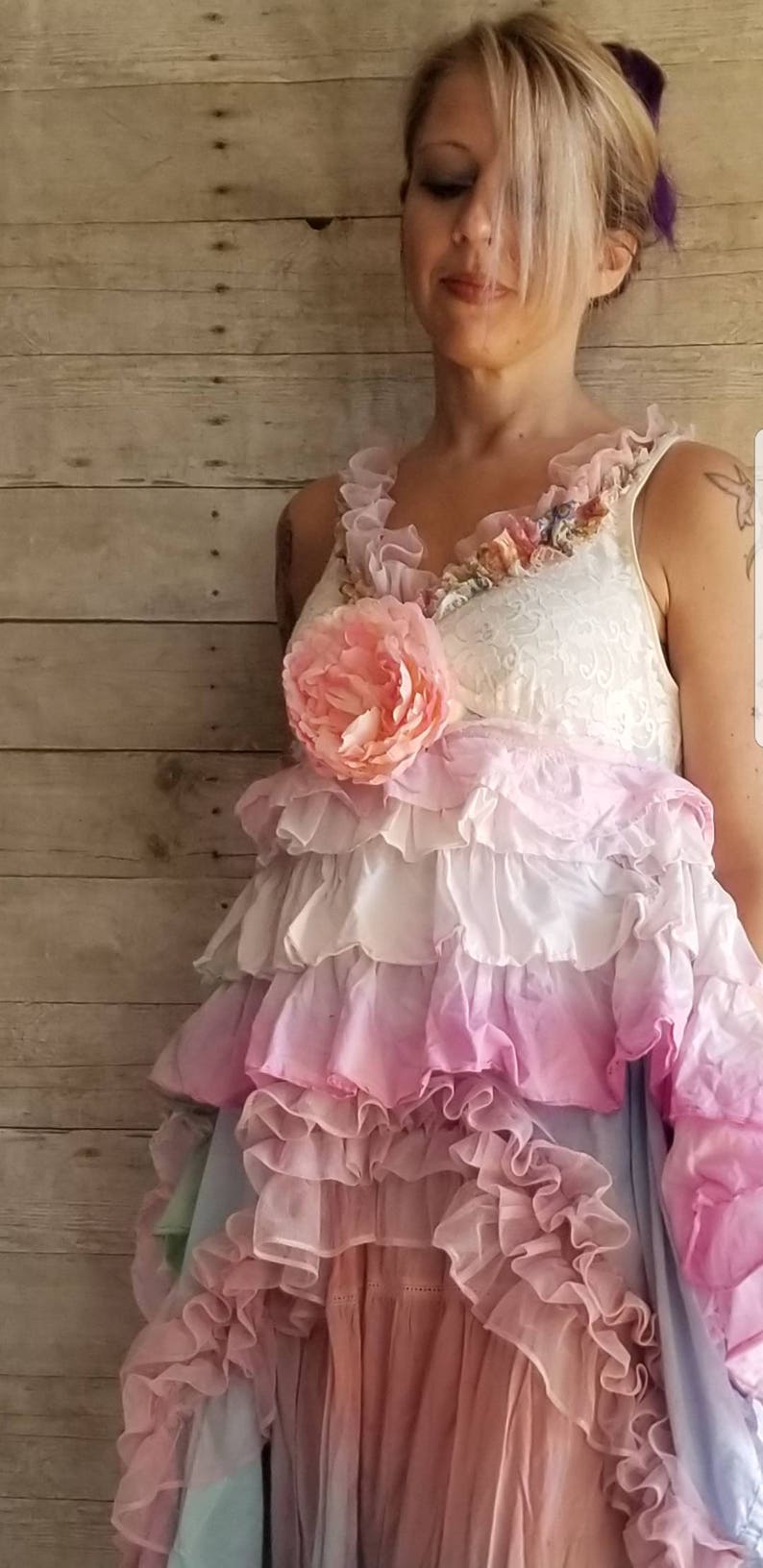 K&ouml;nnte beinhalten: Ein mehrlagiges R&uuml;schenkleid in Rosa-, Wei&szlig;- und Blaut&ouml;nen. Das Kleid hat eine gro&szlig;e, rosa Stoffblume am Oberteil und zarte Spitzendetails. Der Gesamtstil ist romantisch und b&ouml;hmisch.
