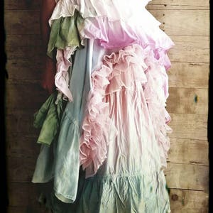 K&ouml;nnte beinhalten: Ein mehrschichtiges Kleid mit R&uuml;schen in Rosa-, Blau- und Gr&uuml;nt&ouml;nen. Das Kleid hat ein Spitzenmieder und eine gro&szlig;e rosa Blume. Der Hintergrund ist eine Holzwand.