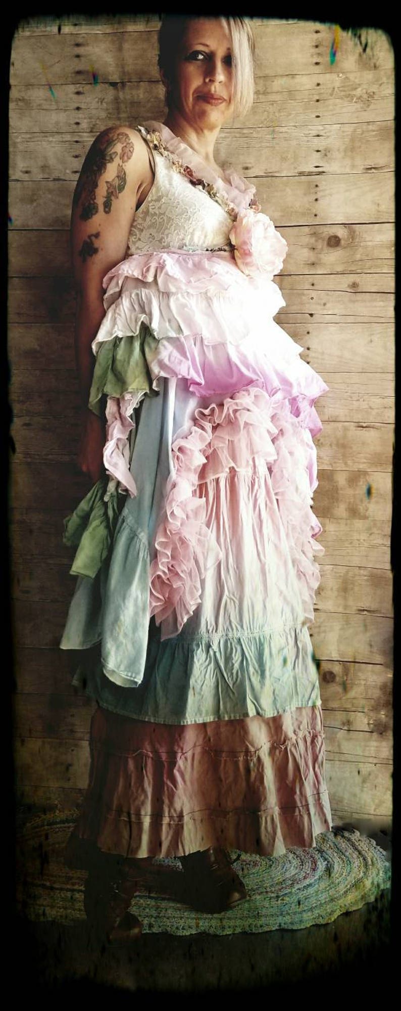 K&ouml;nnte beinhalten: Ein mehrschichtiges Kleid mit R&uuml;schen in Rosa-, Blau- und Gr&uuml;nt&ouml;nen. Das Kleid hat ein Spitzenmieder und eine gro&szlig;e rosa Blume. Der Hintergrund ist eine Holzwand.