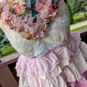 K&ouml;nnte beinhalten: Ein mehrlagiges Kleid mit einem cremefarbenen Spitzenoberteil und einem ger&uuml;schten Rock in Rosa- und Wei&szlig;t&ouml;nen. Der Ausschnitt ist mit einer rosa T&uuml;llr&uuml;sche und floralen Akzenten verziert. Das Kleid hat eine Vintage- und romantische &Auml;sthetik.