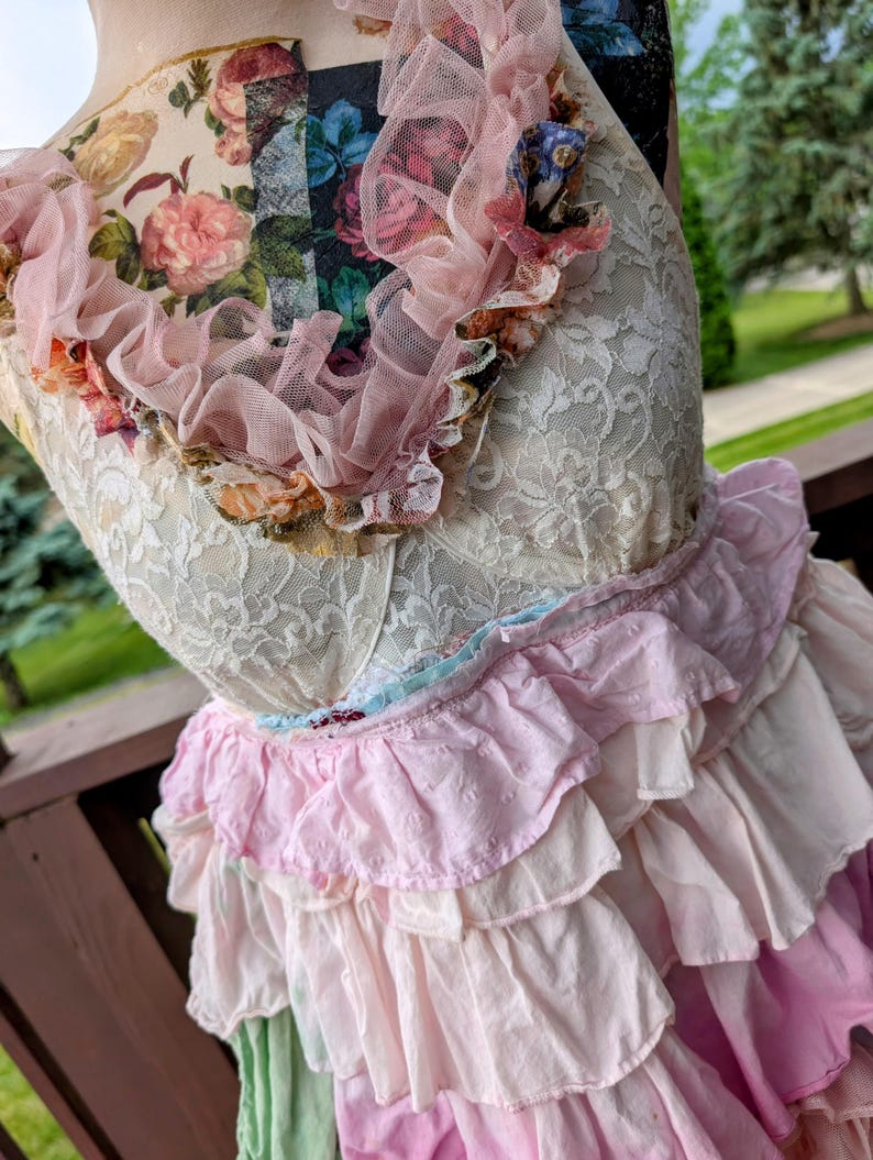 K&ouml;nnte beinhalten: Ein mehrlagiges Kleid mit einem cremefarbenen Spitzenoberteil und einem ger&uuml;schten Rock in Rosa- und Wei&szlig;t&ouml;nen. Der Ausschnitt ist mit einer rosa T&uuml;llr&uuml;sche und floralen Akzenten verziert. Das Kleid hat eine Vintage- und romantische &Auml;sthetik.