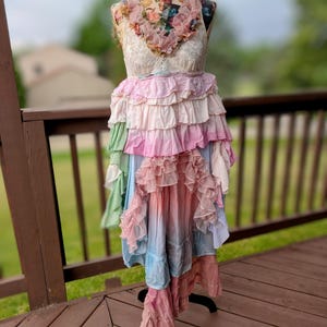 K&ouml;nnte beinhalten: Ein mehrschichtiges R&uuml;schenkleid, das auf einer Schaufensterpuppe pr&auml;sentiert wird. Das Kleid hat ein florales Oberteil, einen Spitzen-Overlay und einen Rock, der von Rosa zu Blau verblasst. Das Kleid ist ein einzigartiger, b&ouml;hmischer Stil.