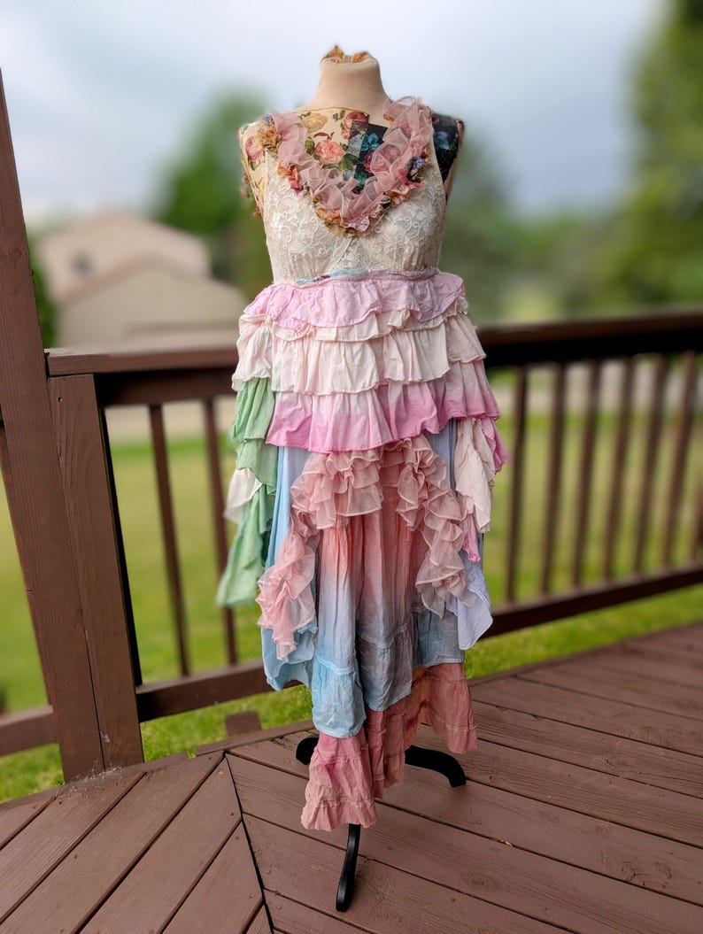 K&ouml;nnte beinhalten: Ein mehrschichtiges R&uuml;schenkleid, das auf einer Schaufensterpuppe pr&auml;sentiert wird. Das Kleid hat ein florales Oberteil, einen Spitzen-Overlay und einen Rock, der von Rosa zu Blau verblasst. Das Kleid ist ein einzigartiger, b&ouml;hmischer Stil.