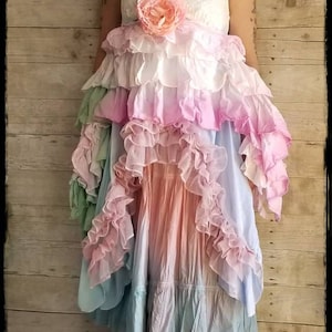 K&ouml;nnte beinhalten: Ein mehrschichtiges Kleid in Pastellt&ouml;nen von Rosa, Blau und Pfirsich. Das Kleid hat eine florale Verzierung am Oberteil und wird mit braunen Stiefeln getragen. Der Hintergrund ist eine Holzwand.