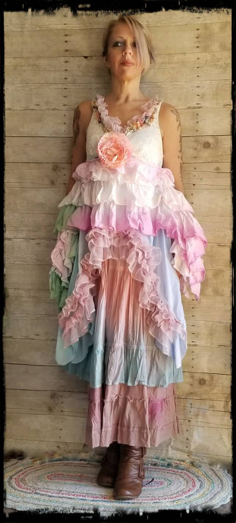 K&ouml;nnte beinhalten: Ein mehrschichtiges Kleid in Pastellt&ouml;nen von Rosa, Blau und Pfirsich. Das Kleid hat eine florale Verzierung am Oberteil und wird mit braunen Stiefeln getragen. Der Hintergrund ist eine Holzwand.