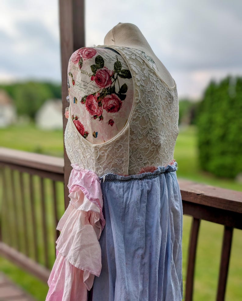 K&ouml;nnte beinhalten: Vintage-Kleid auf einer Schaufensterpuppe. Das Oberteil hat eine cremefarbene Spitze und einen Blumendruck mit rosa Rosen. Der Rock kombiniert hellblauen Stoff und rosa R&uuml;schen, wodurch ein geschichteter Effekt entsteht. Das Kleid wird im Freien ausgestellt.