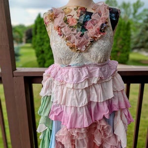 K&ouml;nnte beinhalten: Ein mehrschichtiges Kleid auf einer Schaufensterpuppe. Das Oberteil hat einen Blumendruck und Spitze mit R&uuml;schen am Ausschnitt. Der Rock besteht aus gestuften R&uuml;schen in Rosa, Gr&uuml;n und Blau, was einen Bohemian-Look erzeugt.