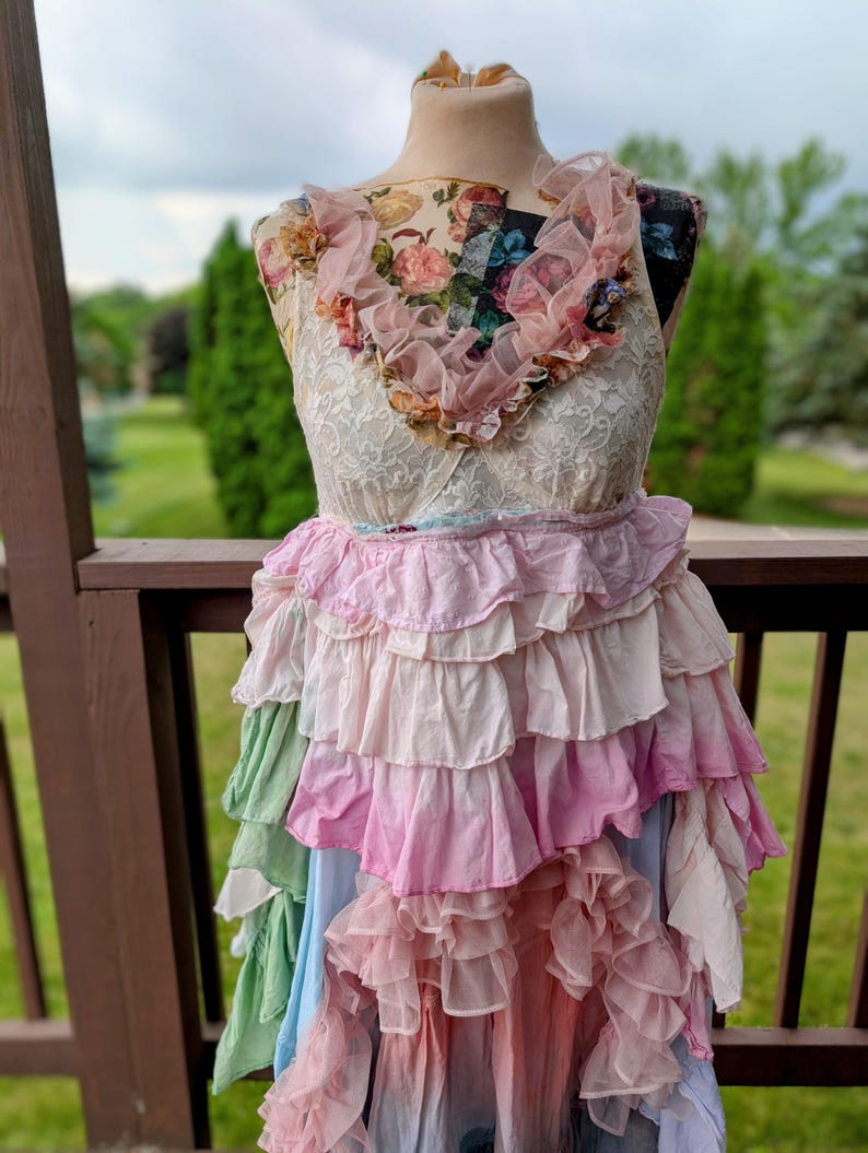 K&ouml;nnte beinhalten: Ein mehrschichtiges Kleid auf einer Schaufensterpuppe. Das Oberteil hat einen Blumendruck und Spitze mit R&uuml;schen am Ausschnitt. Der Rock besteht aus gestuften R&uuml;schen in Rosa, Gr&uuml;n und Blau, was einen Bohemian-Look erzeugt.