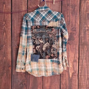 Op de afbeelding: Een teal, wit en tan geruit overhemd met een distressed design. De achterkant heeft een afbeelding van een rodeo-scène met de woorden "Rodeo City". Denim- en kanten patches zijn op het overhemd genaaid.