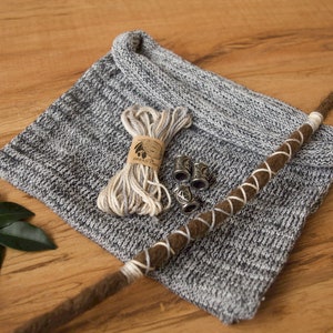 May include: A gray knitted fabric with a brown cord wrapped around it. The cord has white accents and there are five small silver metal pieces on the fabric. There is a spool of white and beige yarn on the fabric.
