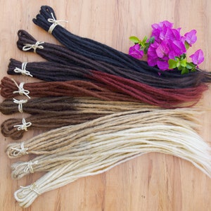 May include: A set of six synthetic dreadlocks in various colors, including black, brown, auburn, and blonde. The dreadlocks are tied together with string and arranged in a fan shape on a wooden surface.